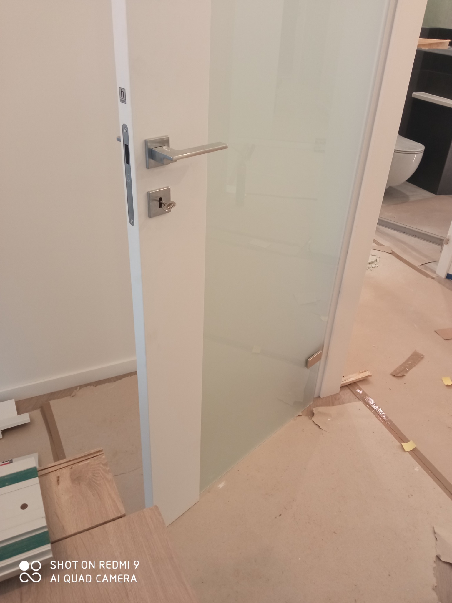 Białe, przeszklone drzwi łazienkowe z srebrną klamką, widoczne panele podłogowe i fragment toalety w tle, pomieszczenie w trakcie prac wykończeniowych.