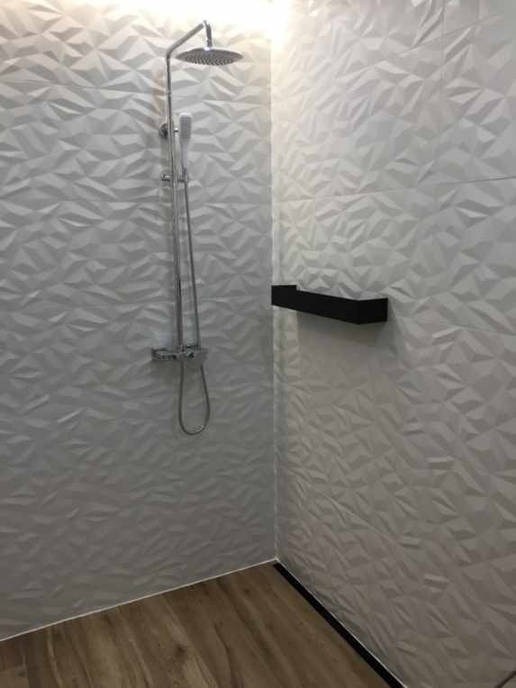 Nowoczesna kabina prysznicowa z trójwymiarowymi, białymi płytkami na ścianach, chromowaną armaturą i drewnopodobną podłogą, uzupełniona o czarną, minimalistyczną półkę.