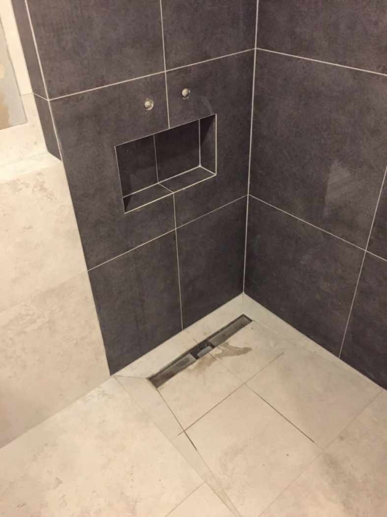 Wykończony fragment łazienki z ciemnymi płytkami na ścianie, wnęką na kosmetyki i odpływem liniowym w podłodze wyłożonej jasnymi płytkami.