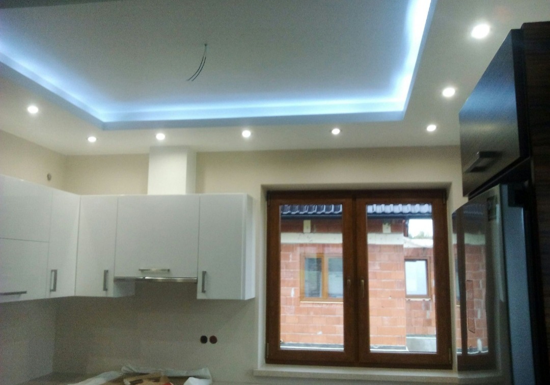 Wykończona kuchnia z białymi szafkami, oświetleniem LED w suficie podwieszanym i brązowymi ramami okiennymi z widokiem na budynek w trakcie budowy.