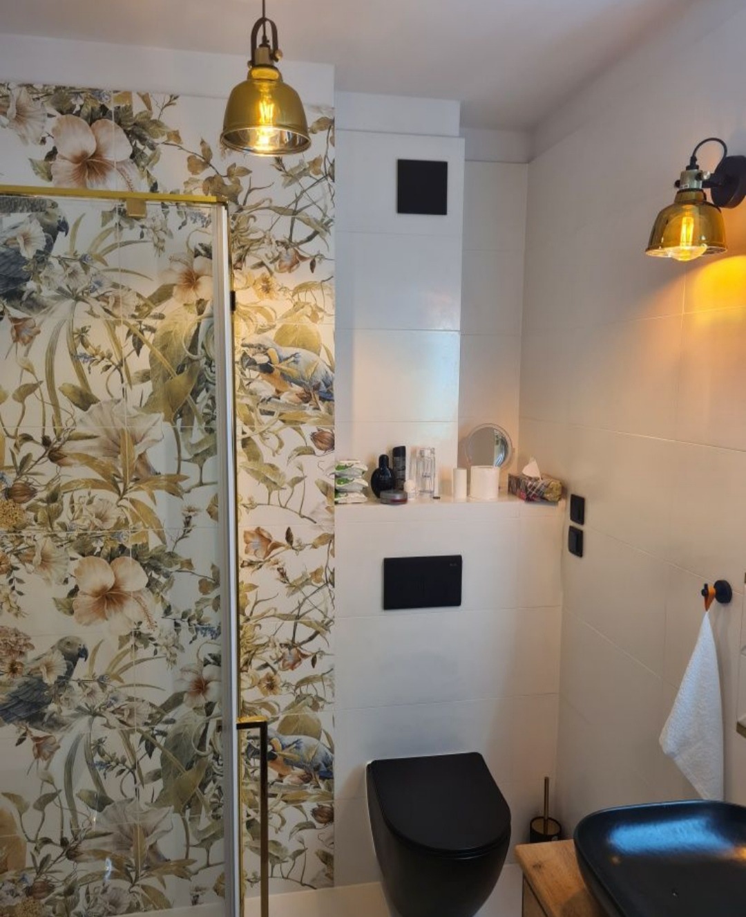 Nowoczesna łazienka z kabiną prysznicową z motywem kwiatowym i ptaków, czarna toaleta i umywalka, oświetlenie w stylu industrialnym z elementami złota, białe płytki na ścianach.