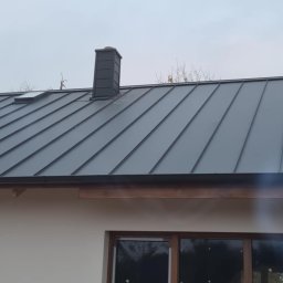 Ag-bud Agnieszka Mikołajczyk - Szary metalowy dach z kominem i oknem dachowym, widok z dołu, w tle pochmurne niebo.