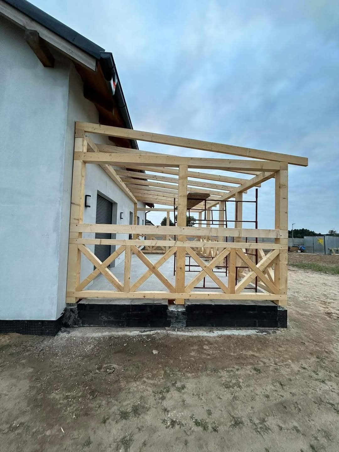 Drewniana konstrukcja zadaszonego tarasu w trakcie budowy, widoczne rusztowania i niezagospodarowany teren wokół budynku.