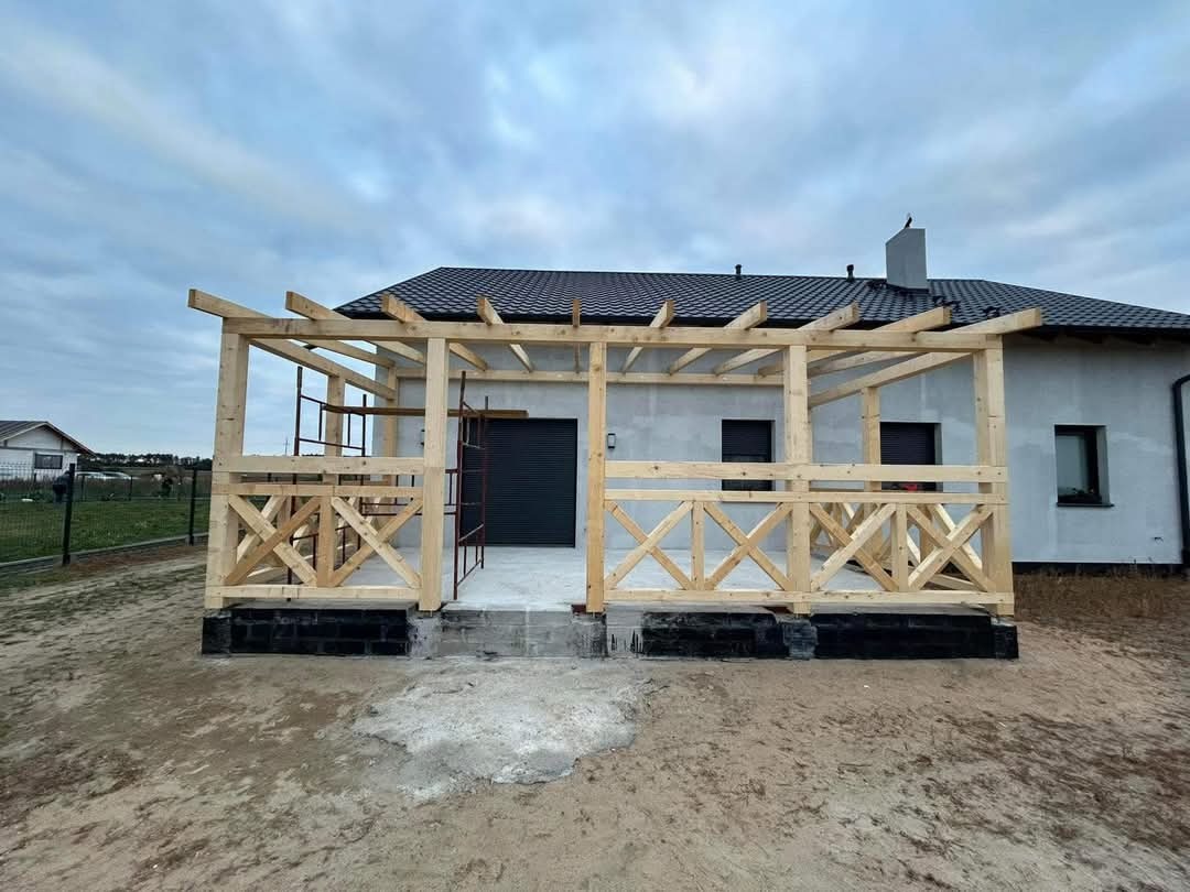 Surowa konstrukcja drewniana werandy z balustradami krzyżowymi, przylegająca do domu z ciemną dachówką i szarą elewacją, widoczne rusztowania i fragmenty betonu na ziemi.