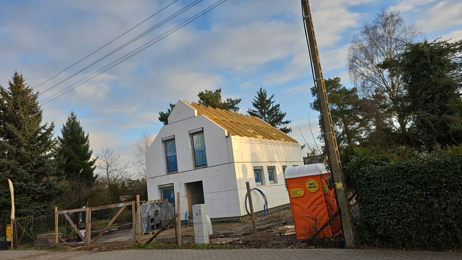 Dom w trakcie budowy z widoczną konstrukcją dachu, styropianem na elewacji i przenośną toaletą WC Skip na posesji.