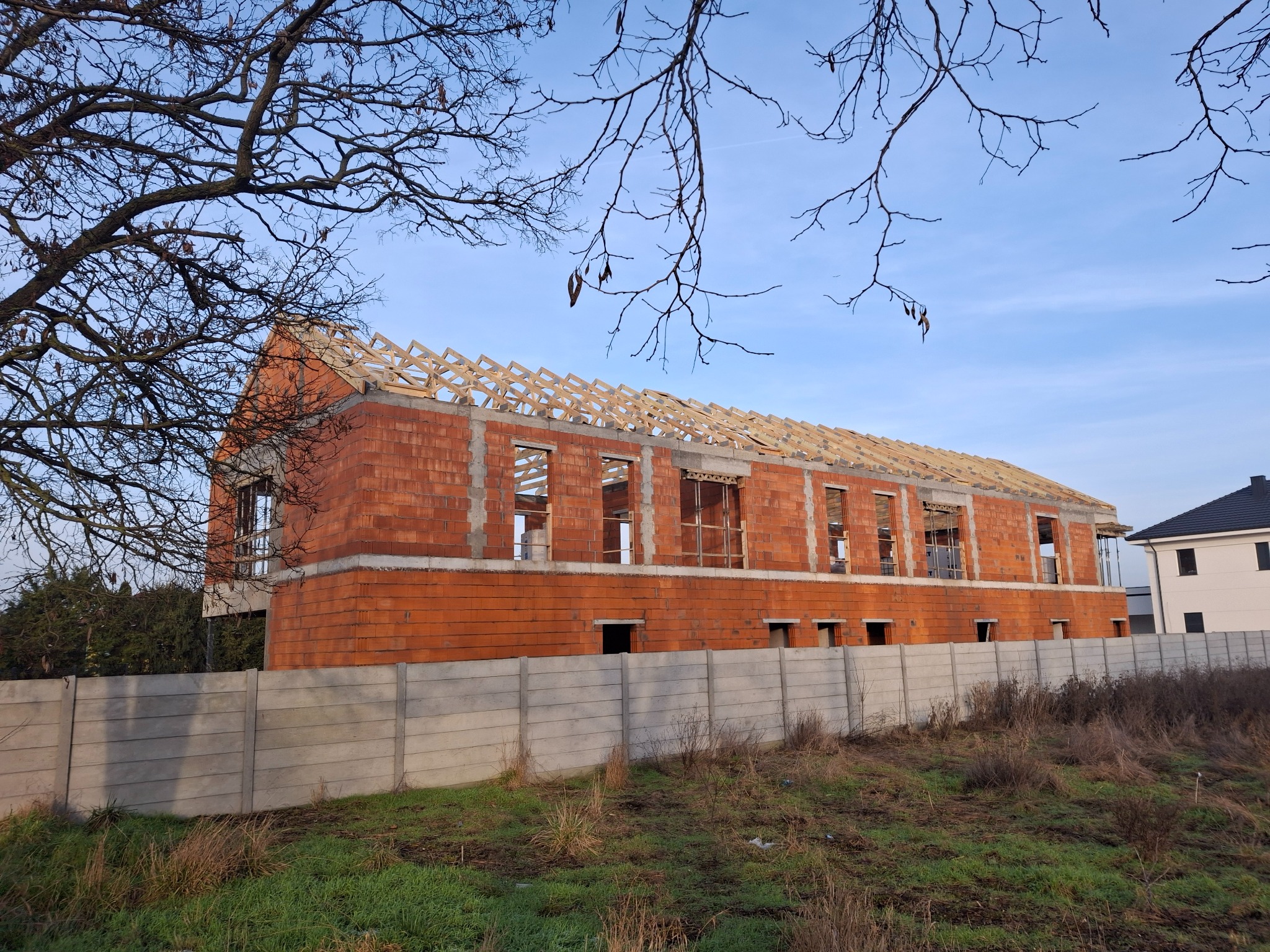 Budynek w trakcie budowy z widoczną konstrukcją drewnianą dachu, ceglanymi ścianami i betonowym ogrodzeniem, sfotografowany w słoneczny dzień z perspektywy niskiego kąta.