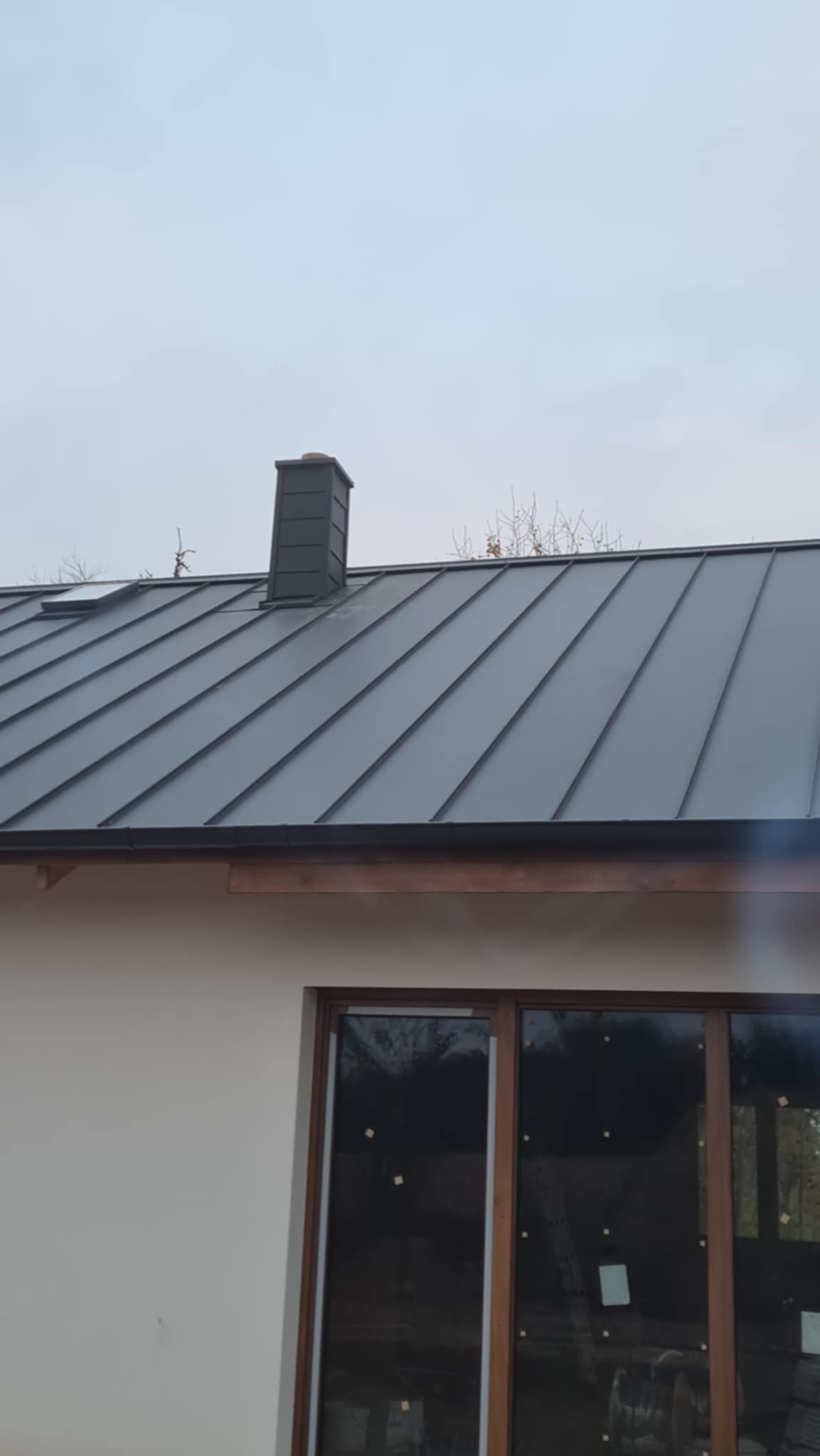 Szary metalowy dach z kominem i oknem dachowym, widok z dołu, w tle pochmurne niebo.
