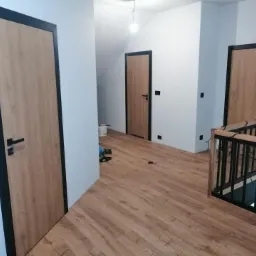 Wnętrze domu po remoncie z trzema zamontowanymi drzwiami wewnętrznymi o drewnopodobnym wykończeniu i czarnych ramach oraz korytarzem wyłożonym panelami imitującymi drewno.