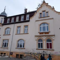 Odnowiona fasada kamienicy z jasną elewacją i białymi oknami o różnych kształtach, widoczne ślady graffiti na dolnej części budynku i przechodzący ludzie.