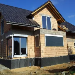 Dom w trakcie budowy z zamontowanymi oknami z białymi ramami i szarą roletą zewnętrzną, widoczne cegły i czarna hydroizolacja fundamentów.