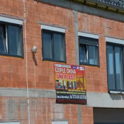 Nowe okna PCV z roletami zewnętrznymi zamontowane w surowym budynku z cegły, widoczny plakat reklamowy producenta okien.