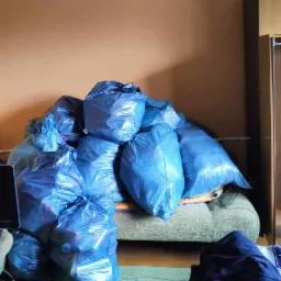 Stos niebieskich worków na śmieci, częściowo wypełnionych, leżących na sofie w pokoju z pomarańczowymi ścianami, przygotowanych do wyniesienia.