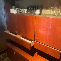 Częściowo opróżniona, brązowa, drewniana komoda z otwartymi szufladami, na górze szkło i naczynia.