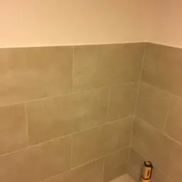 Ukończona ściana łazienki wyłożona beżowymi, matowymi płytkami ceramicznymi, z widoczną lampą roboczą DeWalt w rogu pomieszczenia.