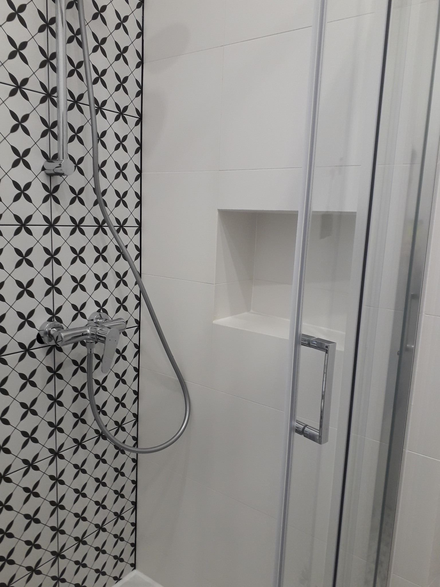 Nowoczesna kabina prysznicowa z chromowaną armaturą i biało-czarną mozaiką na ścianie, wnęka w ścianie wyłożona białymi płytkami, szklane drzwi przesuwne z metalowym uchwytem.