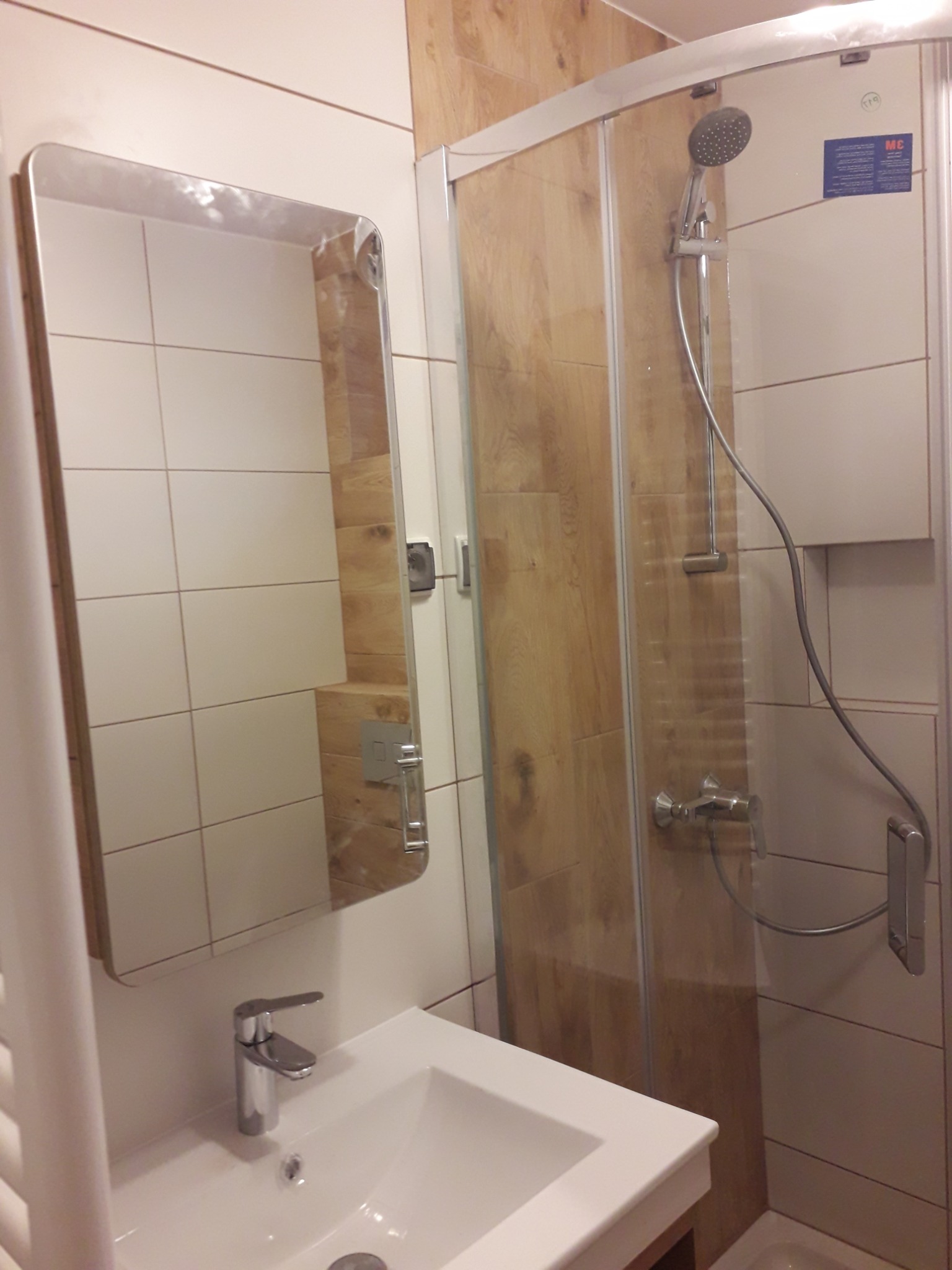 Nowoczesna łazienka z kabiną prysznicową z panelami imitującymi drewno, umywalką nablatową i dużym lustrem z zaokrąglonymi rogami.