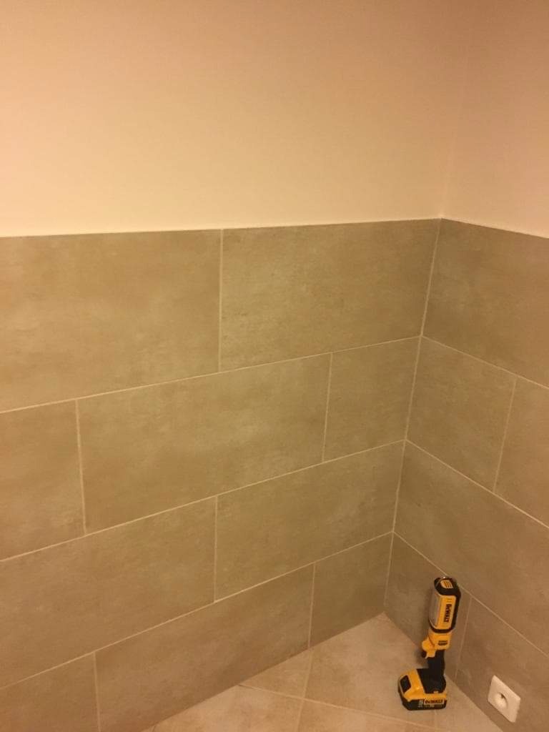 Ukończona ściana łazienki wyłożona beżowymi, matowymi płytkami ceramicznymi, z widoczną lampą roboczą DeWalt w rogu pomieszczenia.