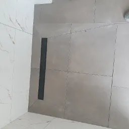 Ułożone szare płytki podłogowe z odpływem liniowym w kabinie prysznicowej, widoczny gumowy młotek i fragment buta, białe płytki imitujące marmur na ścianie.