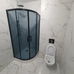 Nowoczesna łazienka z kabiną prysznicową z czarnymi ramami i marmurowymi płytkami na ścianach i podłodze, widoczna toaleta z otwartą klapą.