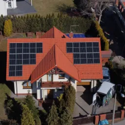 Widok z góry na dom z czerwoną dachówką, na której zamontowane są panele słoneczne; obok domu zaparkowany biały bus z otwartymi drzwiami, a w tle widoczne budynki w trakcie budowy.