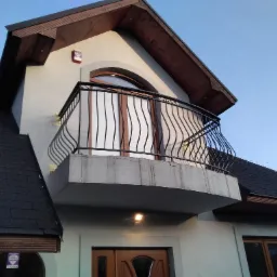 Czarna, metalowa balustrada balkonowa z dekoracyjnymi, falistymi prętami pionowymi, zamontowana na betonowym balkonie nowoczesnego domu z brązową stolarką okienną i dachem z ciemnej dachówki...