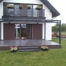 Nowoczesny taras z szarych płytek z czarną metalową konstrukcją zadaszenia, donice z kwiatami, balkon z balustradą na piętrze.