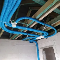 Niebieskie rury wentylacyjne, zamocowane pod drewnianą konstrukcją dachu w budynku w Cedrach Wielkich, z widocznymi białymi kształtkami i fragmentem ściany z białych bloczków.