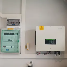 Biała skrzynka z bezpiecznikami i licznikami na ścianie obok białego inwertera fotowoltaicznego Sofar z wyświetlaczem, okablowanie widoczne, gniazdko elektryczne w pobliżu.