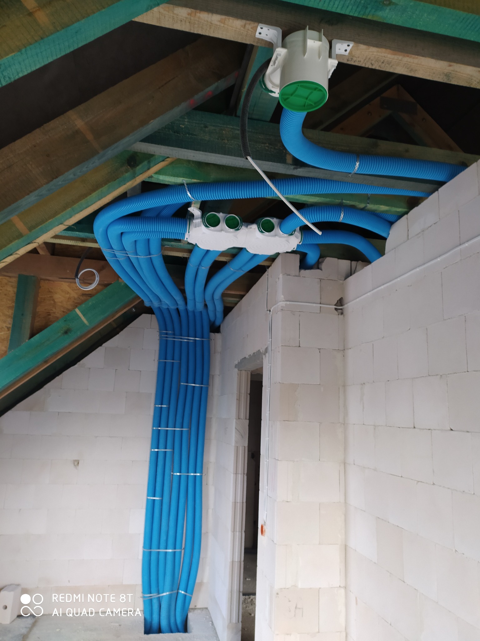 Niebieskie rury wentylacyjne prowadzone wzdłuż ściany z bloczków, rozprowadzone pod drewnianą konstrukcją dachu. Centrala wentylacyjna z zielonymi elementami widoczna pod sufitem.