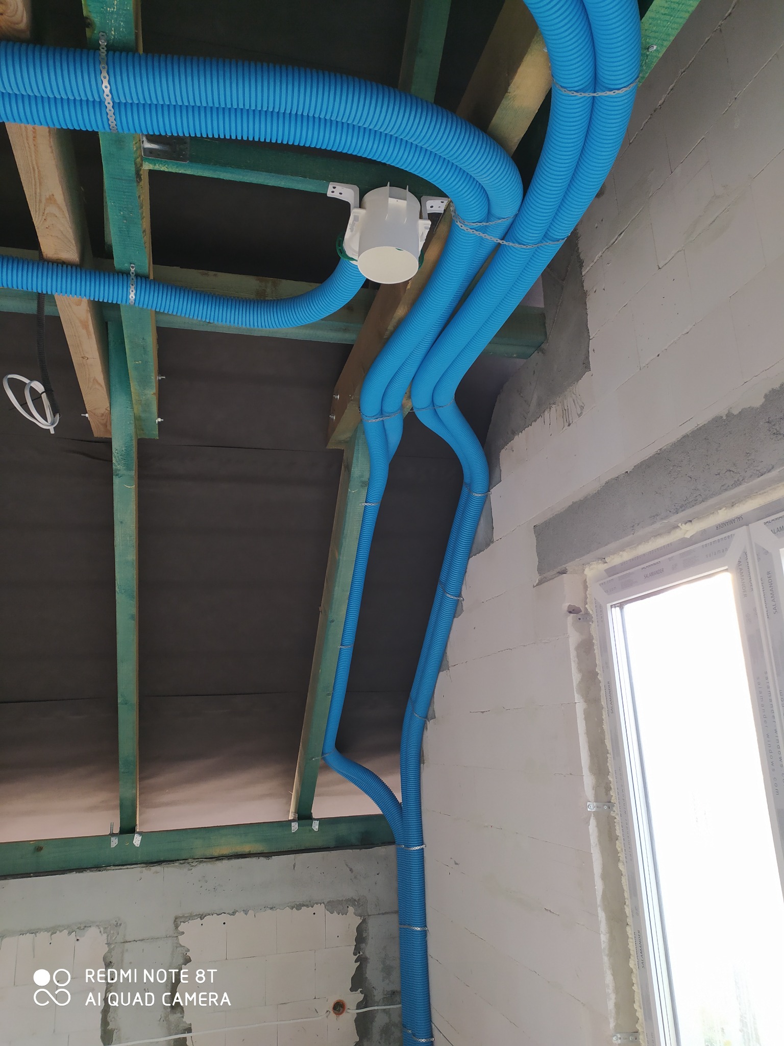 Niebieskie rury wentylacyjne korugowane, zamocowane metalowymi opaskami do drewnianej konstrukcji stropu w surowym wnętrzu budynku w trakcie budowy.