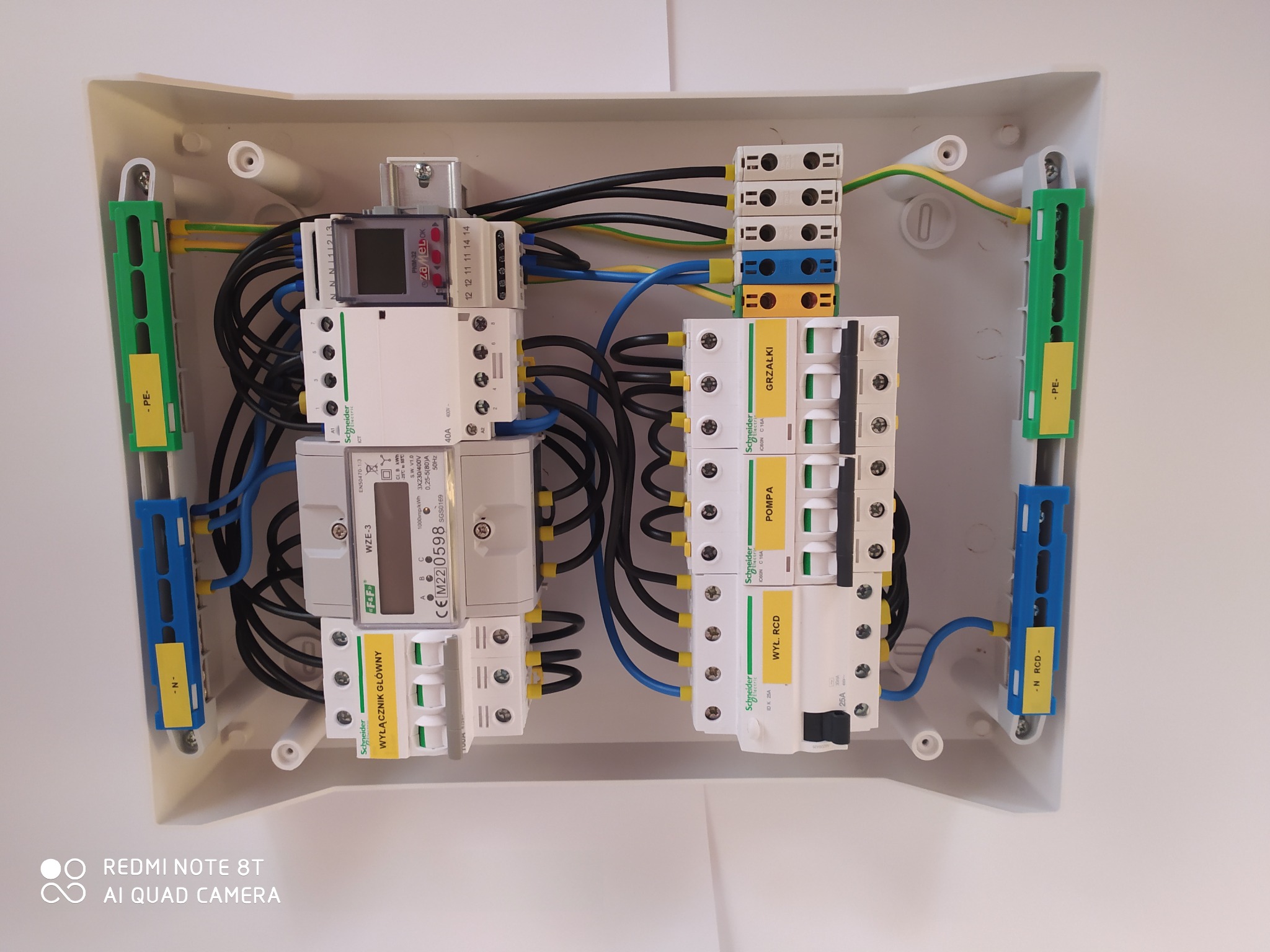 Rozdzielnica elektryczna w białej obudowie, widok z góry, z zainstalowanymi bezpiecznikami, wyłącznikiem głównym, licznikiem energii elektrycznej oraz przewodami w kolorach czarnym, niebieskim...