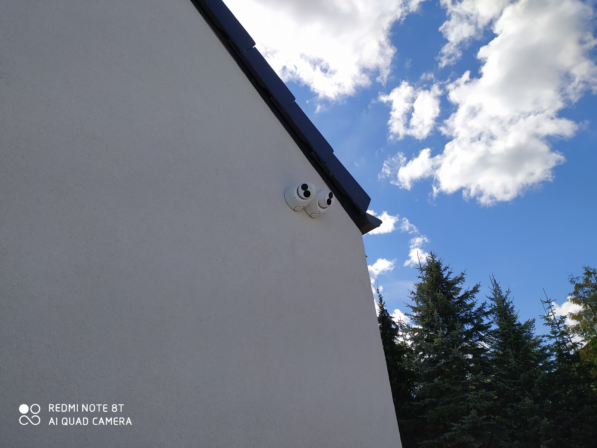 Dwie białe kamery monitoringu zamontowane na elewacji budynku, widok z dołu na tle błękitnego nieba z chmurami i fragmentów zielonych drzew.