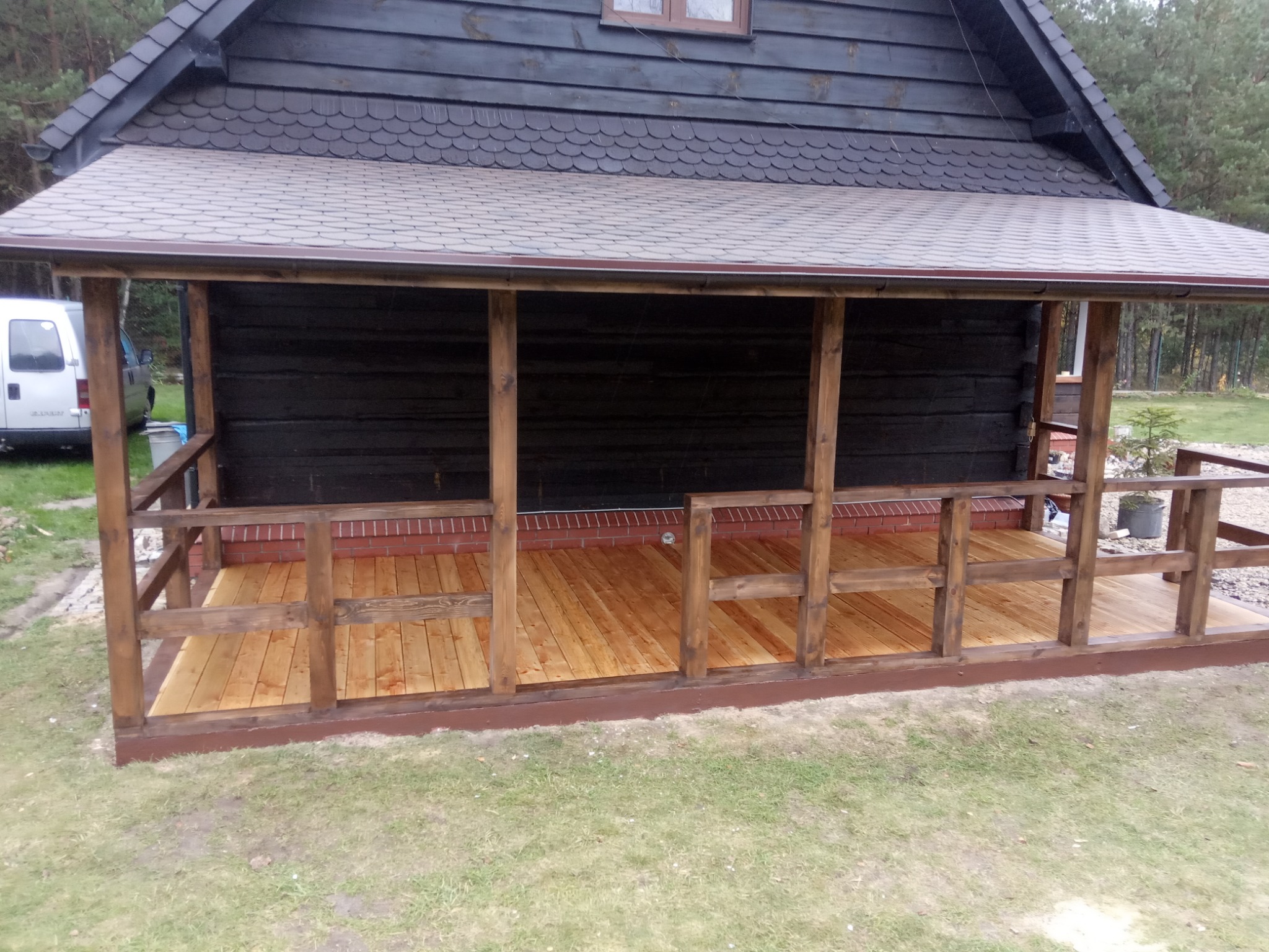 Zadaszony taras z drewnianą podłogą i balustradą, widoczny fragment ściany budynku obitego ciemnym drewnem i fragment białego vana w tle.