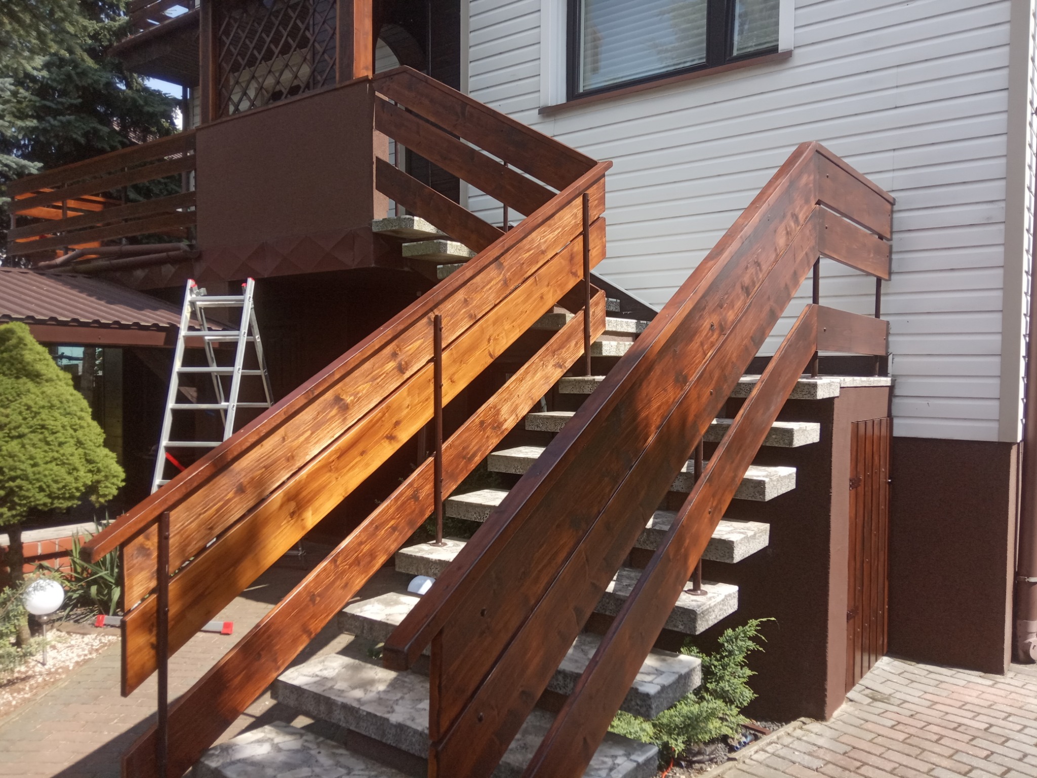 Zewnętrzne schody z drewnianą poręczą w kolorze ciemnego orzecha, prowadzące do wejścia budynku z sidingiem; w tle drabina oparta o balkon.