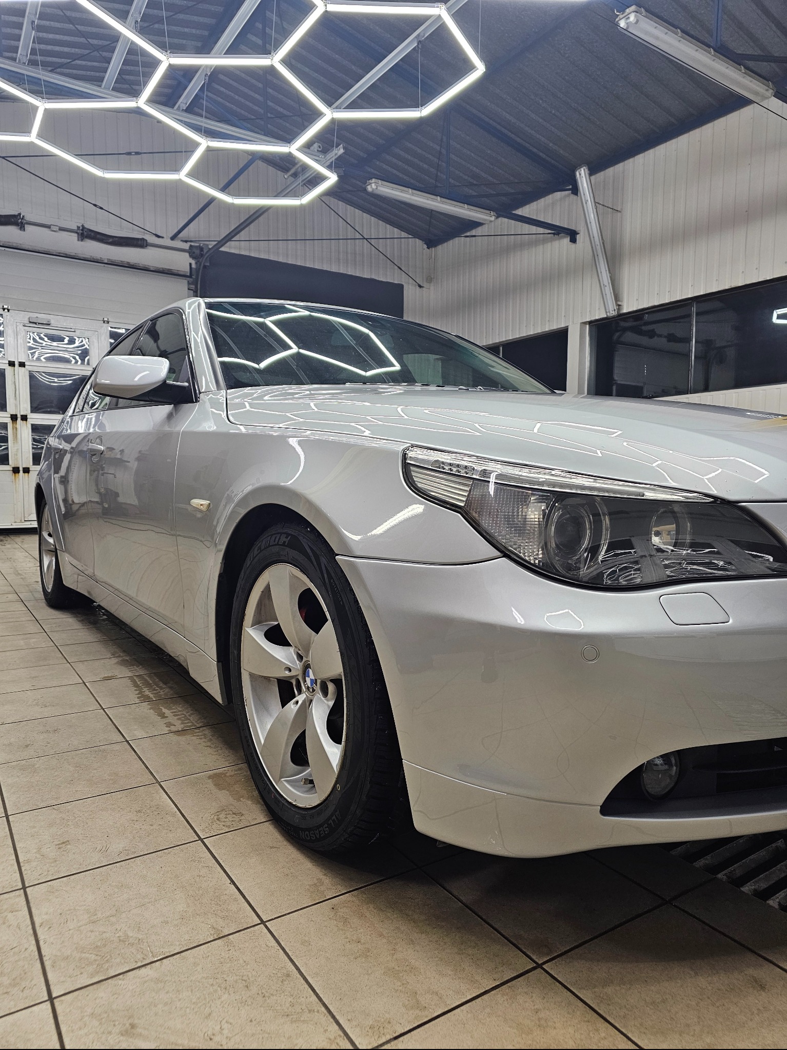 Błyszczący, srebrny samochód BMW po umyciu w myjni, widoczny z niskiej perspektywy, z heksagonalnymi lampami LED na suficie. Czyste felgi i karoseria.