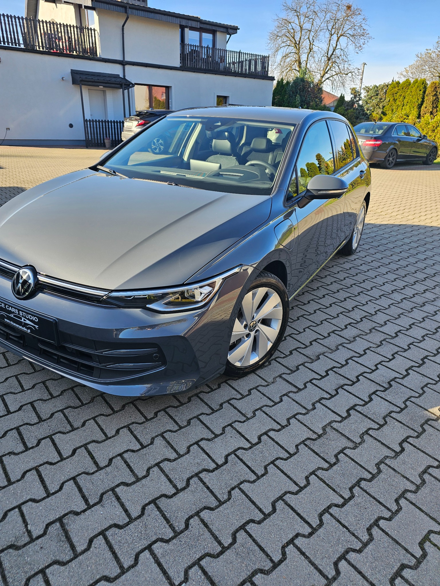 Szary Volkswagen Golf po detailingu, z widocznym logo Nice Cars Studio na tablicy rejestracyjnej, zaparkowany na kostce brukowej przed budynkiem.