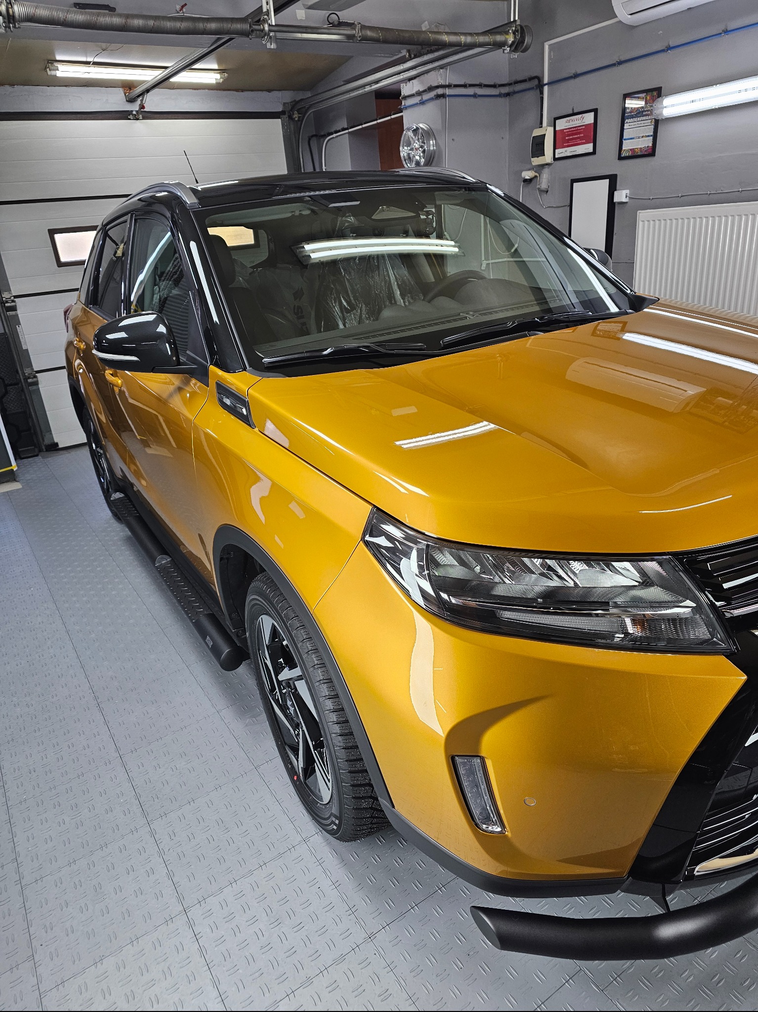 Żółty SUV Suzuki Vitara po oklejeniu folią w garażu, widok z przodu na reflektor i błotnik. Błyszcząca powierzchnia, czarne dodatki. Podłoga garażu z szarej kostki.