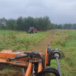Widok z perspektywy operatora na pomarańczową kosiarkę bijakową w trakcie pracy na zarośniętym polu, z widocznym czerwonym traktorem w tle i świeżo skoszonym pasem trawy.