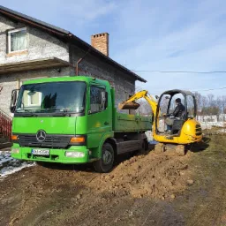 Żółta mini koparka załadowuje ziemię na zieloną ciężarówkę na błotnistym terenie przed domem.