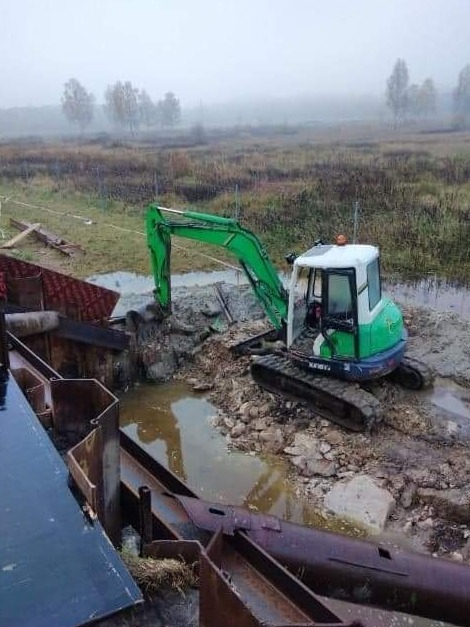 Zielona mini koparka w błotnistym terenie, obok widoczne metalowe elementy konstrukcyjne, prawdopodobnie zabezpieczenie wykopu. Mglista pogoda w tle.