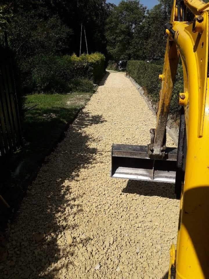 Żółta mini koparka z łyżką rozkłada jasny żwir na ścieżce wzdłuż żywopłotu w słoneczny dzień.