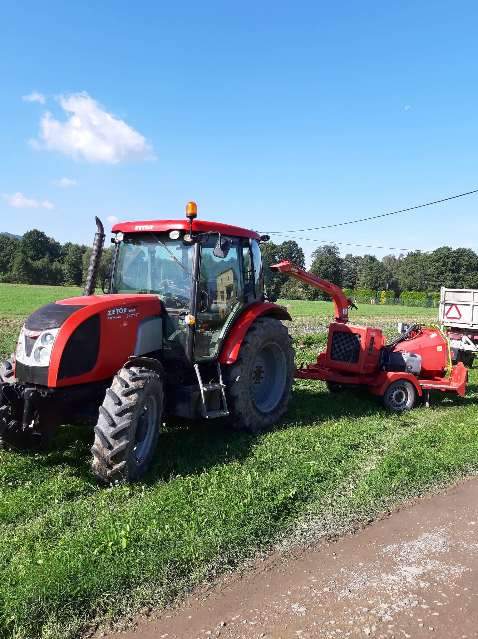 Czerwony traktor Zetor z przyczepioną rębarką do gałęzi na zielonym polu, widoczne drzewa w tle i błękitne niebo z chmurami.