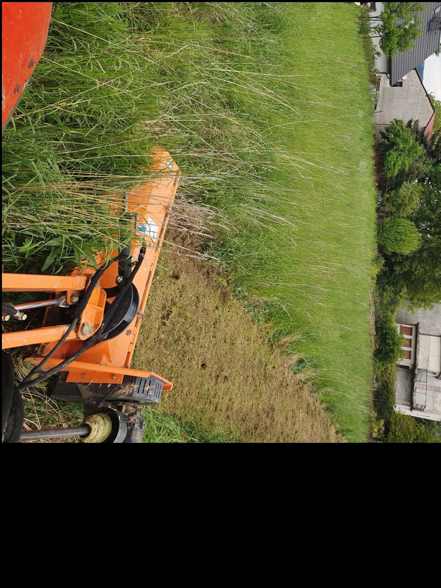 Pomarańczowa kosiarka bijakowa w trakcie koszenia wysokiej trawy na działce, z widocznym fragmentem budynku w tle.