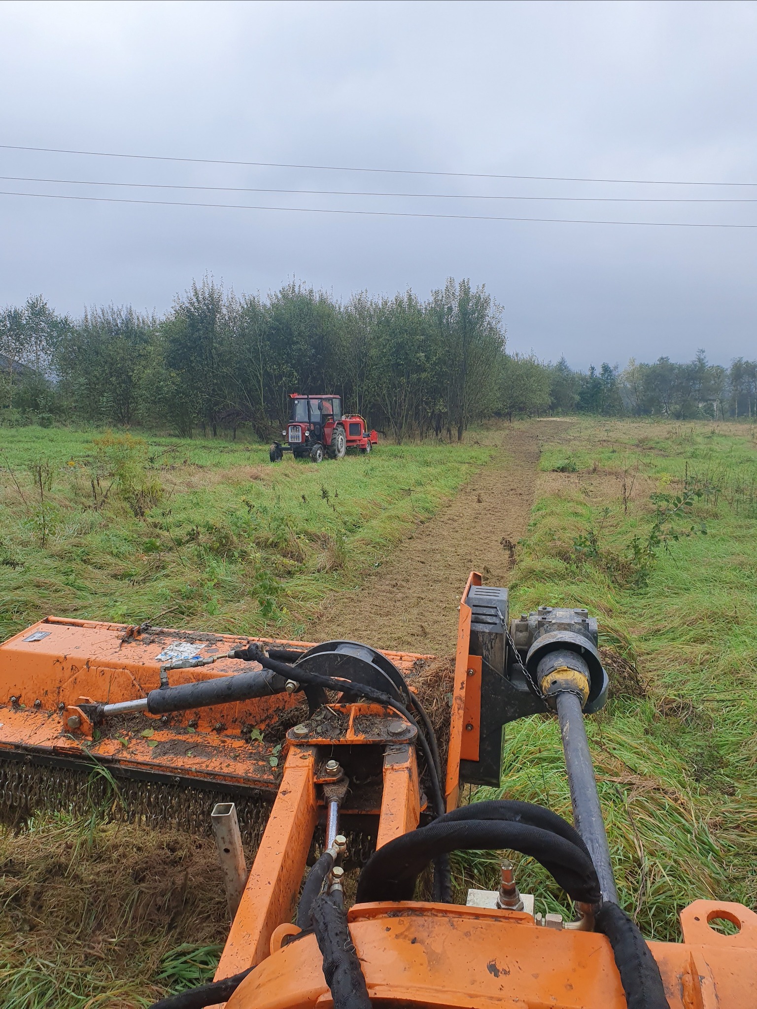 Widok z perspektywy operatora na pomarańczową kosiarkę bijakową w trakcie pracy na zarośniętym polu, z widocznym czerwonym traktorem w tle i świeżo skoszonym pasem trawy.