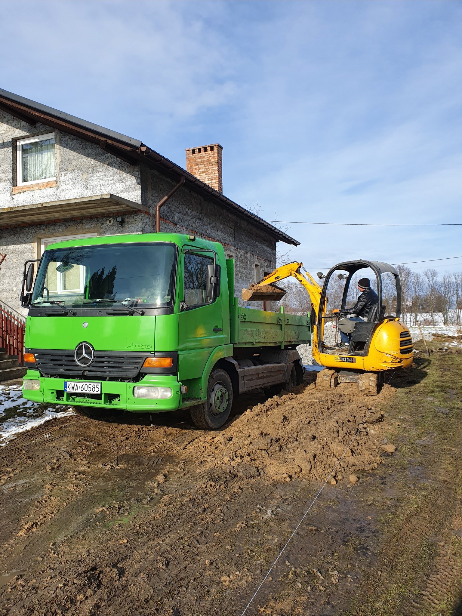 Żółta mini koparka załadowuje ziemię na zieloną ciężarówkę na błotnistym terenie przed domem.