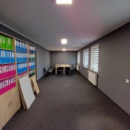 Firma Remontowo - Budowlana Damian Nowakowski - Wnętrze biura po malowaniu: szare ściany, ciemna wykładzina, regał z segregatorami, stół z krzesłami. Widoczne grzejniki pod oknami z żaluzjami.