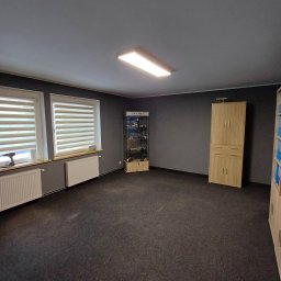 Firma Remontowo - Budowlana Damian Nowakowski - Pomalowane na szaro wnętrze pokoju z oknami z żaluzjami, grzejnikami, szafką ekspozycyjną i szafą. Na parapecie wiertarka akumulatorowa.