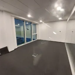 Puste, jasne biuro z dużymi oknami i szarą wykładziną, widoczne gniazda podłogowe i białe ściany.