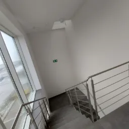 Klatka schodowa w nowoczesnym budynku z szarymi płytkami na schodach i metalową balustradą, widok z góry w kierunku okna i znaku ewakuacyjnego.