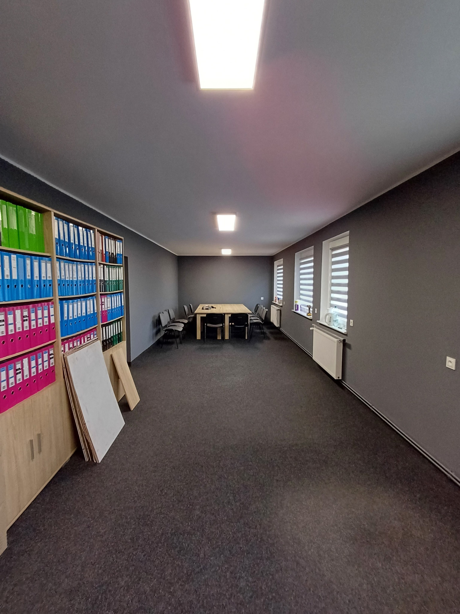 Wnętrze biura po malowaniu: szare ściany, ciemna wykładzina, regał z segregatorami, stół z krzesłami. Widoczne grzejniki pod oknami z żaluzjami.